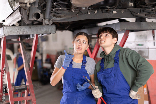 Obraz na płótnie Two focused young auto mechanics, girl and guy, in blue overalls working together, inspecting car lifted on hydraulic lift in vehicle repair shop and discussing reason for breakdown w salonie