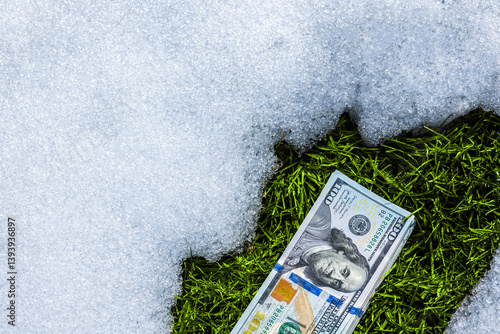Stack of us dollar banknotes on the snow near thawed green grass patch