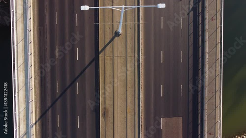 Person walks past fencing at Budapest rail station during dusk with city blur in background, drone top down static as tram crosses bridge by cars