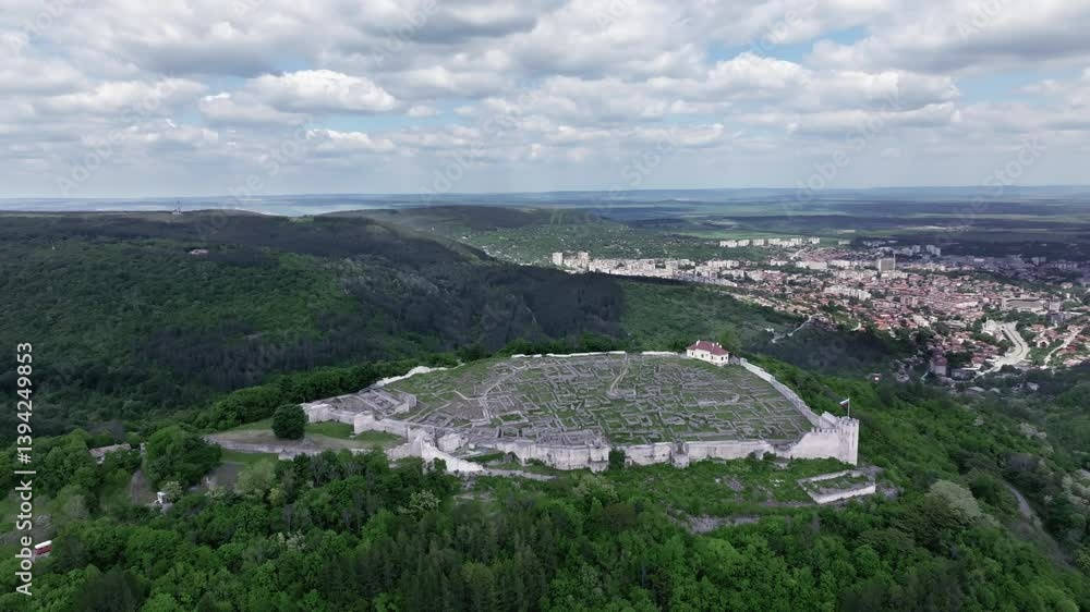 Aerial view of the ancient Shumen Fortress surrounded by green forested hills, with the modern town of Shumen in the background under a partly cloudy sky. Aerial view of the ancient Shumen Fortress surrounded by green forested hills, with the modern town of Shumen in the background under a partly cloudy sky.