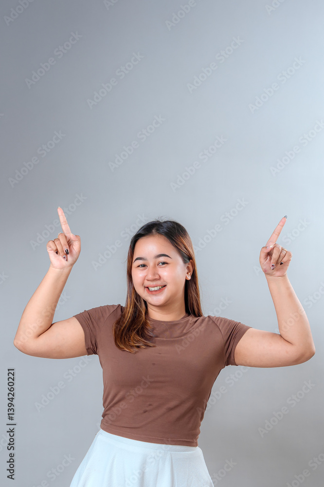 Cheerful young woman smiling brightly while pointing upwards with both index fingers, showing excitement and positivity. Perfect for promotion, advertisement, or copy space designs. Cheerful young woman smiling brightly while pointing upwards with both index fingers, showing excitement and positivity. Perfect for promotion, advertisement, or copy space designs.