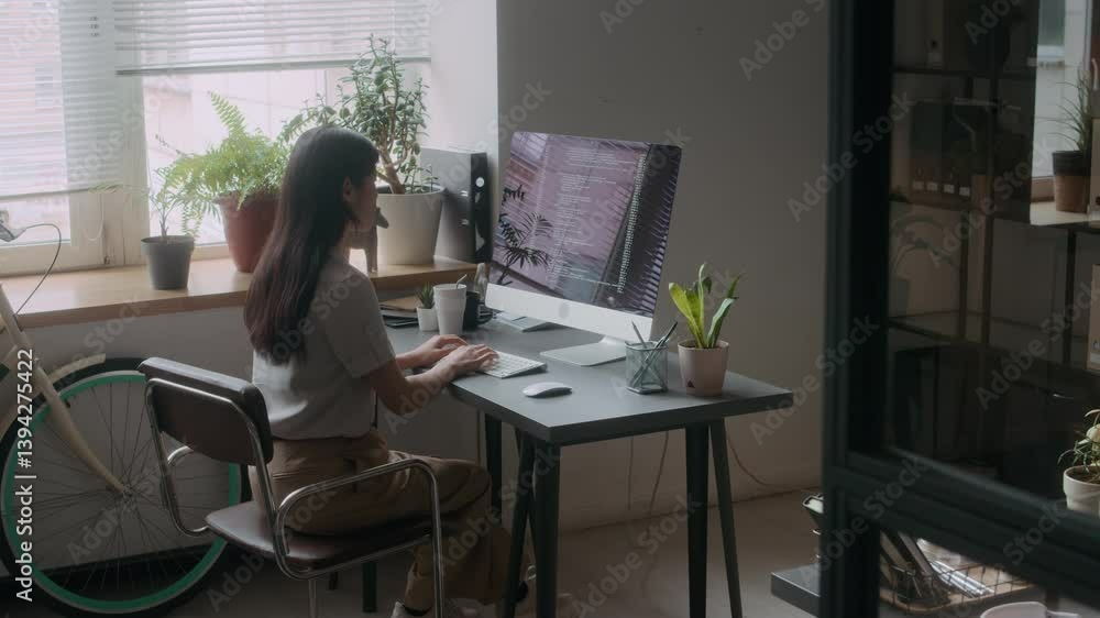 Young female programmer typing code on desktop computer in modern comfortable office with plants and bicycle by window