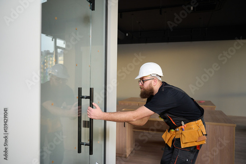 Bearded worker installing glass door in the new office. Construction worker installing door in a new house construction site. Builder in uniform adjusting glass door in new apartment.