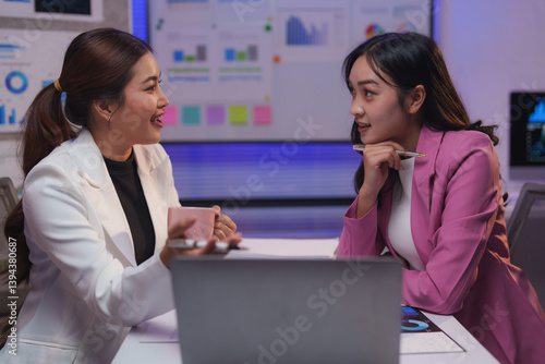 Two asian businesswomen are discussing strategy in a modern office, using a laptop and analyzing charts and graphs displayed on a large screen, while enjoying a cup of coffee