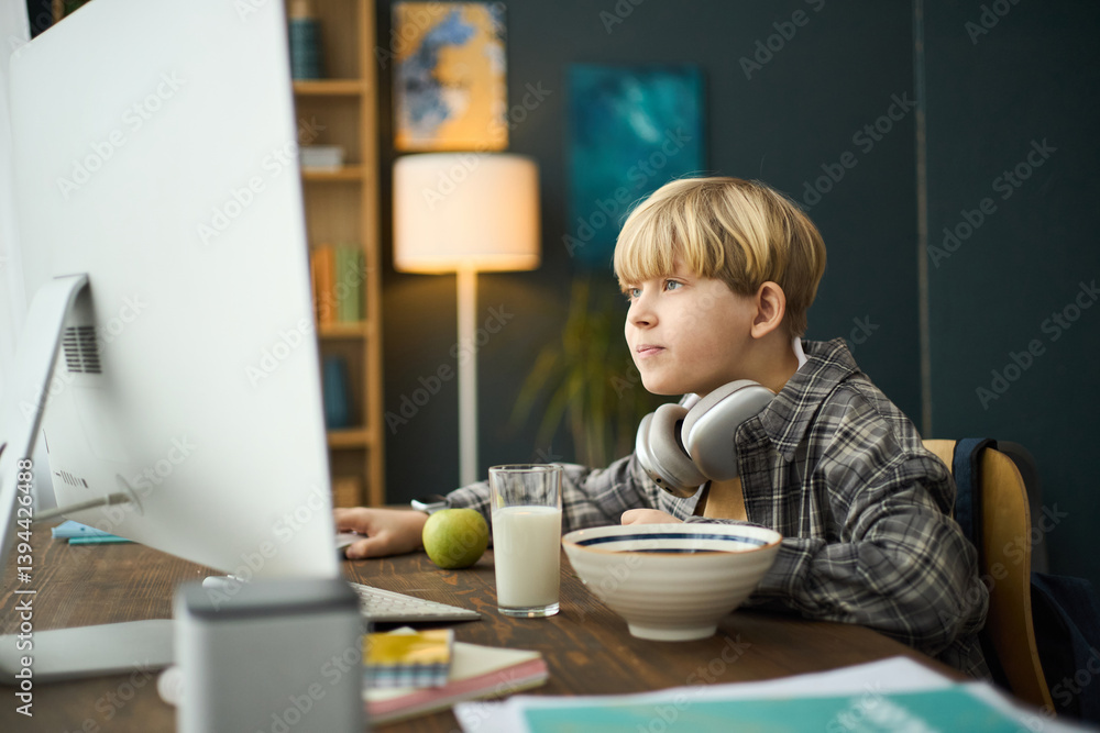 Young Caucasian boy in plaid shirt sitting at desk, watching computer screen attentively with headphones on neck, with apple, glass of milk, and bowl Young Caucasian boy in plaid shirt sitting at desk, watching computer screen attentively with headphones on neck, with apple, glass of milk, and bowl