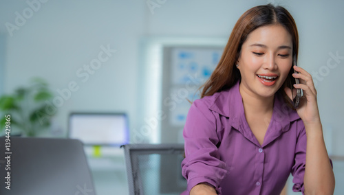 Free Download Young Asian businesswoman talking on phone and smiling while working with laptop in modern office, professional female employee using smartphone for business communication - GFXLoad