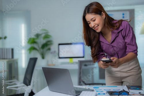 Young Asian businesswoman smiling while using a smartphone and working on a laptop in a modern office during a break from her professional duties