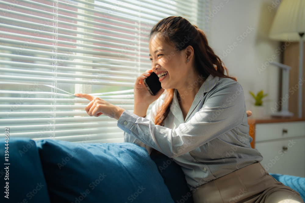 Obraz premium Businesswoman sitting on a sofa, chatting on her phone while pointing at the window blinds, soaking in the natural light and enjoying a bright view of the sunny day outside