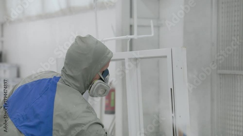A fully equipped worker in protective gear applies a smooth white coating to a window frame using a spray gun in a clean industrial paint booth environment