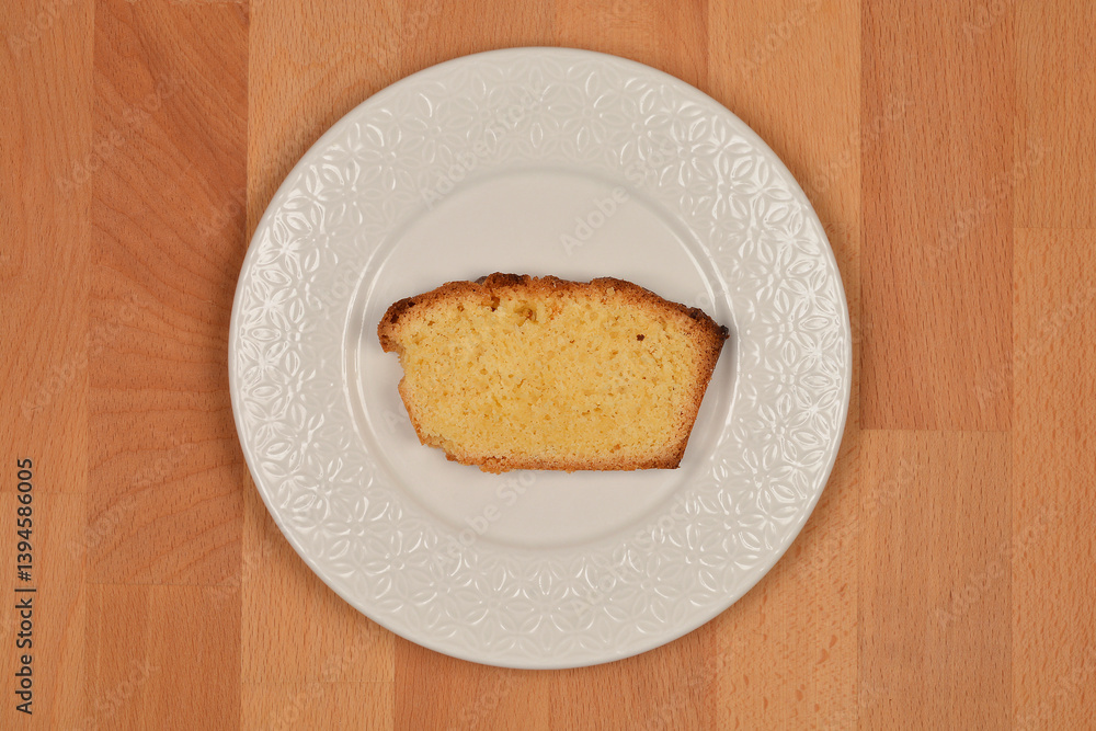 A slice of lemon pound cake is centered on a white decorative plate, which rests on a light brown wooden surface A slice of lemon pound cake is centered on a white decorative plate, which rests on a light brown wooden surface