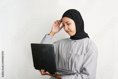 Frustrated young Asian woman holding a laptop while touching her head, showing signs of stress, confusion, or mental fatigue, standing against a plain background with a worried expression.