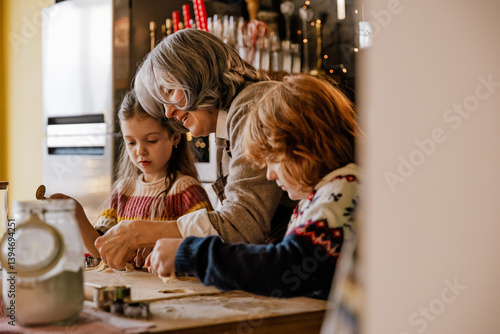A woman is baking cookies with a young girl and a red-haired boy in a cozy kitchen. The children are focused on the dough as they cook together during a holiday at home.