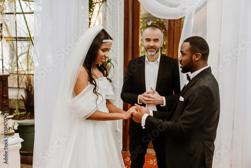 A Black groom and a bride with long dark hair wearing a white wedding dress and veil hold hands during a wedding ceremony indoors while a White male officiant stands between them.