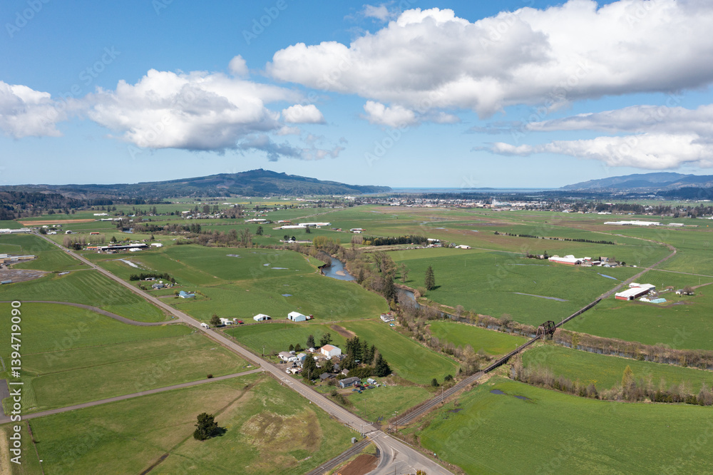 Fototapeta premium Aerial views of lush green countryside in Tillamook, Oregon, a small coastal town near the Pacific Ocean, showing farmlands, country roads, rural houses, mountains, and blue sky in spring-summer