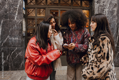 Φωτογραφία On the street, against the building, a young Black woman checks her phone as three female friends gather around, looking at the screen