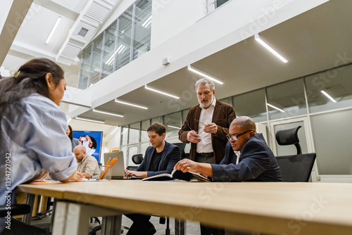 Wallpaper Mural A 55-year-old White businessman dressed in a brown business suit, standing next to the table of his colleagues in an office room, as they're having a meeting, and reviewing some papers of his coworker Torontodigital.ca
