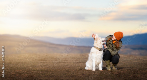 A cheerful young woman in outdoor clothing next to her golden retriever dog on a mountain meadow during sunset. Travel with dog. Life with dog.