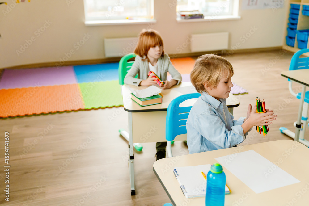 Obraz premium Two White school-aged boys sit at separate desks in a sunlit light classroom with windows and a playmat, the foreground blonde boy and the background red-haired boy sit with colored pencils.