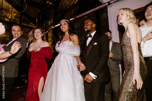A Black bride in her 20s wearing a white dress holds hands with her Black groom in his 20s wearing a black tuxedo, surrounded by multiethnic guests, all dancing, at a wedding in a greenhouse at night.