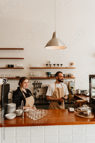 Two baristas standing behind the counter of an empty, cozy caf?, an overweight White woman in her 20s, and a mid-30s White man, smiling and looking to the side while wearing aprons over their clothes