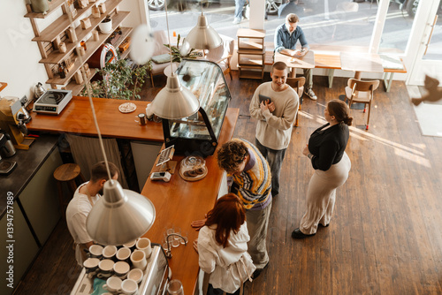 Canvas Print From a high angle inside a sunlit cafe, casually dressed White adult customers and workers stand talking at the wooden counter, while a White man in his 40s sits at a table using his phone nearby