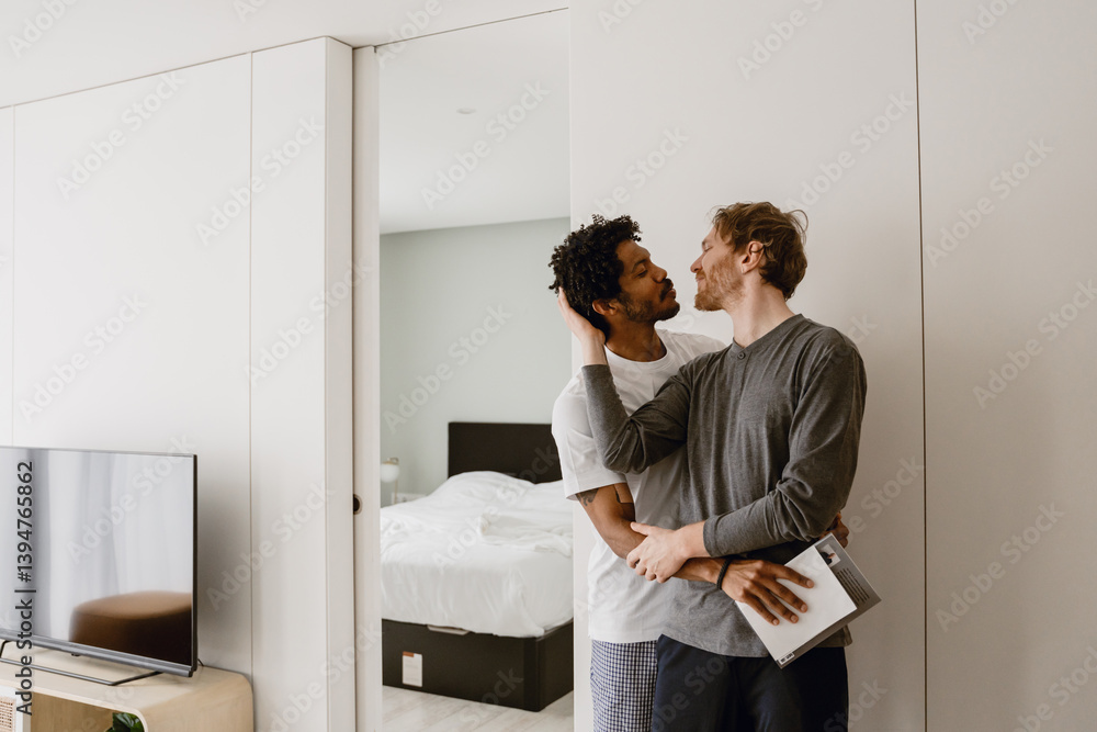 A Caucasian man aged 35 who is dressed in a gray long-sleeve, standing at home in a minimalistic white apartment and leaning back to kiss his mid-30s Black husband, who stands behind him