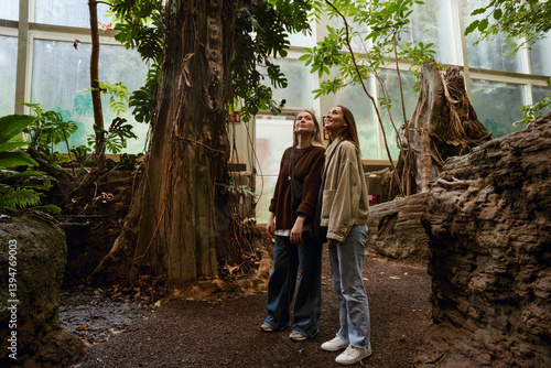 Two young White women with long hair stand in a greenhouse surrounded by tropical plants and trees. One wears a brown sweater, and the other a beige jacket. They admire the natural scenery.