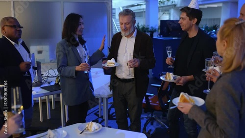 A White elder man is listening to his Asian female colleague's toast, while holding a piece of cake and a glass of champagne, standing among a group of his colleagues during his retirement party.