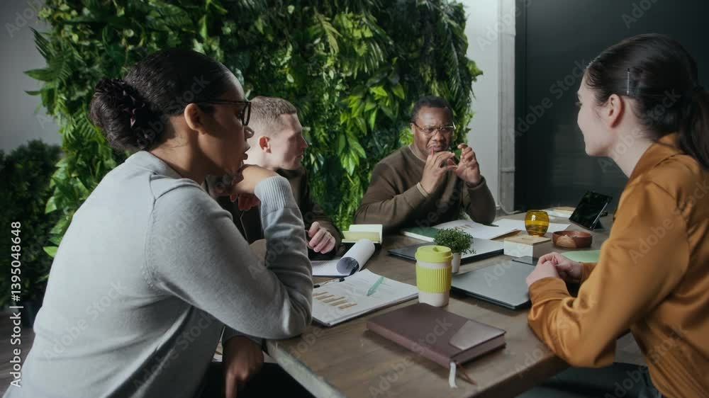 Medium shot of young inspired Black man sharing ideas during brainstorm meeting in modern green office with young multiethnic colleagues sitting at table and collaborating together Medium shot of young inspired Black man sharing ideas during brainstorm meeting in modern green office with young multiethnic colleagues sitting at table and collaborating together