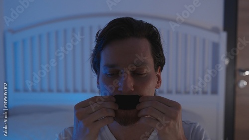 Man in Bed Applying Black Mouth Tape Over Lips with Both Hands Under Dim Blue Light, Anti Snoring and Nose Breathing Concept