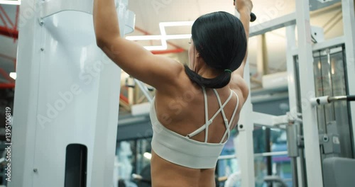 Back view of athletic girl in white top doing back muscle exercises on block trainer in modern gym. Back muscle development and training in the gym