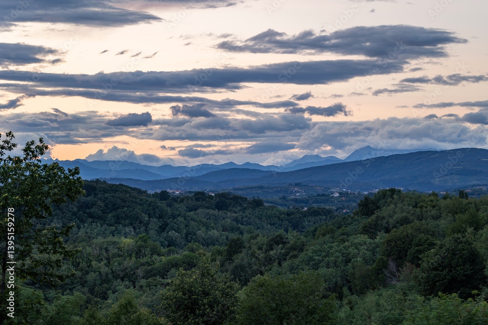 vasto ambiente collinare naturale che si estende fino alle catene montuose delle Alpi lungo il confine tra Italia e Slovenia, al tramonto, in estate, con cielo nuvoloso