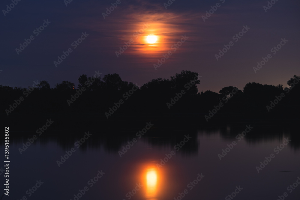 luna piena e di colore arancione che si specchia di notte sull'acqua tranquilla di una palude fluviale, in un ambiente naturale con alberi e cielo leggermente nuvoloso, in estate