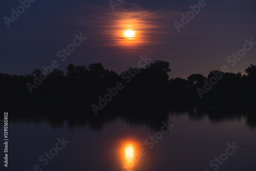 Obraz na płótnie luna piena e di colore arancione che si specchia di notte sull'acqua tranquilla di una palude fluviale, in un ambiente naturale con alberi e cielo leggermente nuvoloso, in estate w salonie