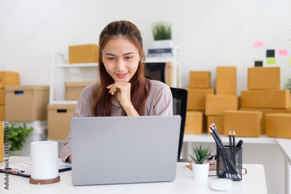 Startup business owner using laptop at desk laptop with packaging boxes in the background. Small business operations and e-commerc concept. Startup business owner using laptop at desk laptop with packaging boxes in the background. Small business operations and e-commerc concept.