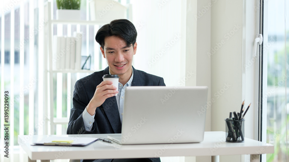 Young Asian businessman smiling while holding a cup of coffee and working on a laptop at a modern office desk. Young Asian businessman smiling while holding a cup of coffee and working on a laptop at a modern office desk.