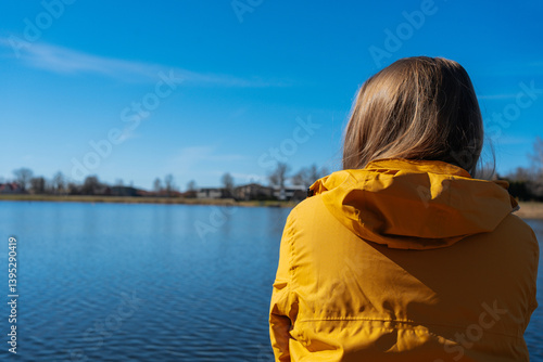 Photos A young woman in a yellow jacket stands on the shore of a lake or river, gazing into the distance