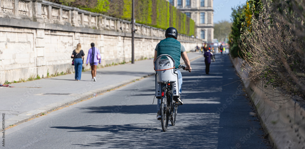 Obraz premium Un homme avec un casque en train de faire du vélo à Paris, sur une piste cyclable. Sur son vélo, il y a un siège bébé. En arrière plan, un bâtiment du Louvre. Le format de l'image est horizontal.