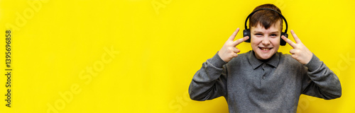A ten year old boy in a gray knitted sweater enthusiastically listens to music in black headphones and shows a gesture with two fingers on a yellow background.