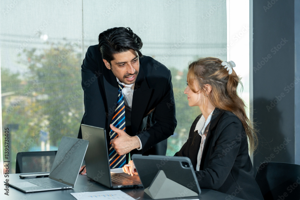 Middle Eastern businessman leaning forward pointing to laptop screen while talking to Caucasian female colleague sitting at office desk with digital tablet and paper during business discussion