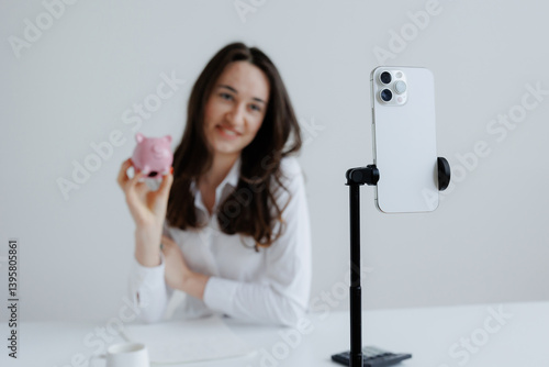 Young woman engages in online content creation while holding a pink piggy bank in a bright, minimalist workspace
