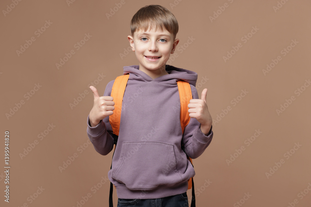 Cute schoolboy with backpack showing thumbs up on brown background