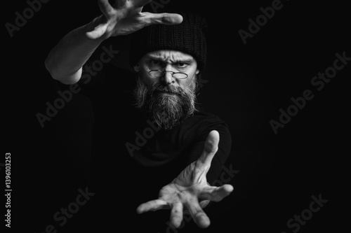 Obraz na płótnie Black and white, dramatic portrait of an energetic man with a beard and glasses, reaching towards the camera. Subtle lighting, emotional expression, dark background, mysterious concept w salonie