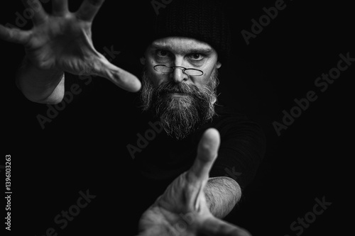 Obraz na płótnie Black and white, dramatic portrait of an energetic man with a beard and glasses, reaching towards the camera. Subtle lighting, emotional expression, dark background, mysterious concept w salonie