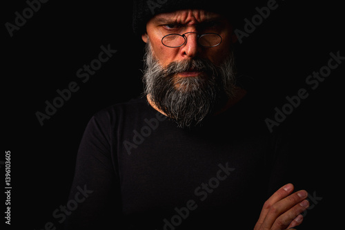 Obraz na płótnie Portrait of a confident mature man with arms crossed in front of him, wearing a knitted hat and glasses. Staring gaze, gray beard and black vest. Stylish discreet portrait on a dark background w salonie