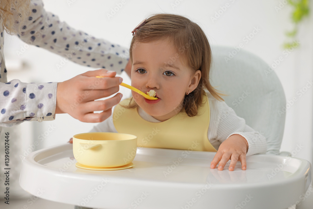 Mother feeding her cute little baby in high chair indoors, closeup Mother feeding her cute little baby in high chair indoors, closeup