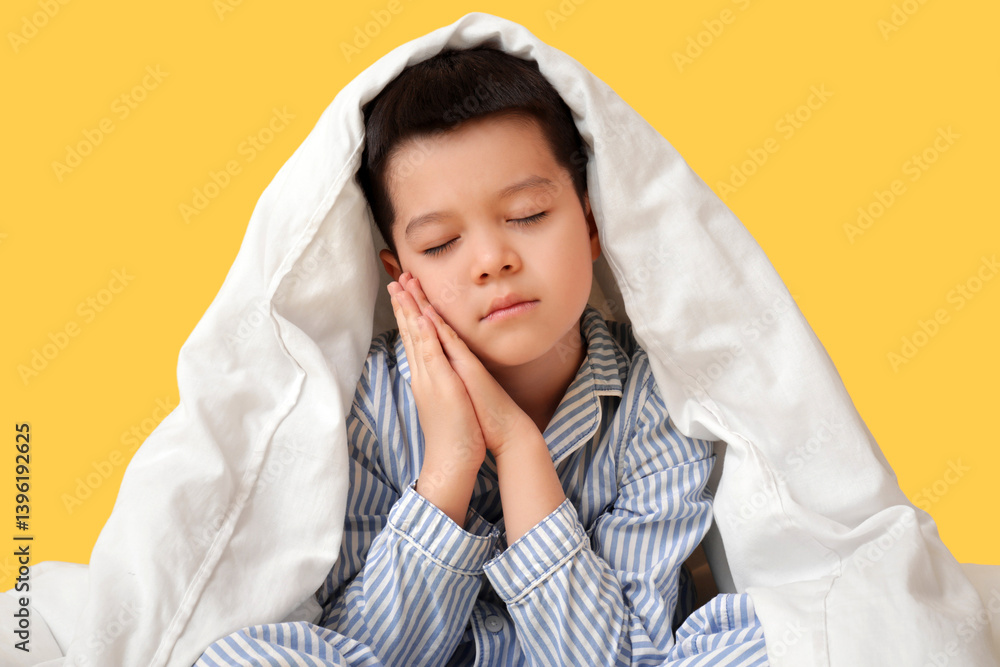 Sleepy little Asian boy in pajamas with blanket sitting on yellow background