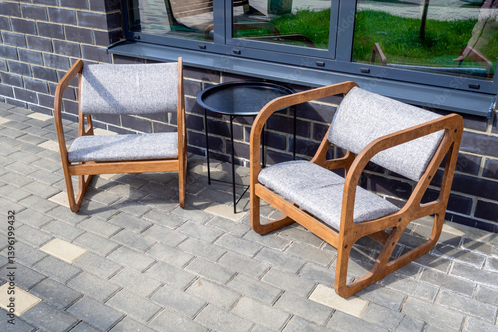 Two wooden armchairs or lounge chairs with cushions and round metal table set on paved sidewalk. Intended for outdoor cafes or relaxation zones Two wooden armchairs or lounge chairs with cushions and round metal table set on paved sidewalk. Intended for outdoor cafes or relaxation zones