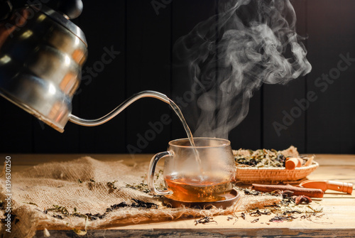Pour hot tea into cups, herbal tea glasses are arranged on a rustic wooden table.