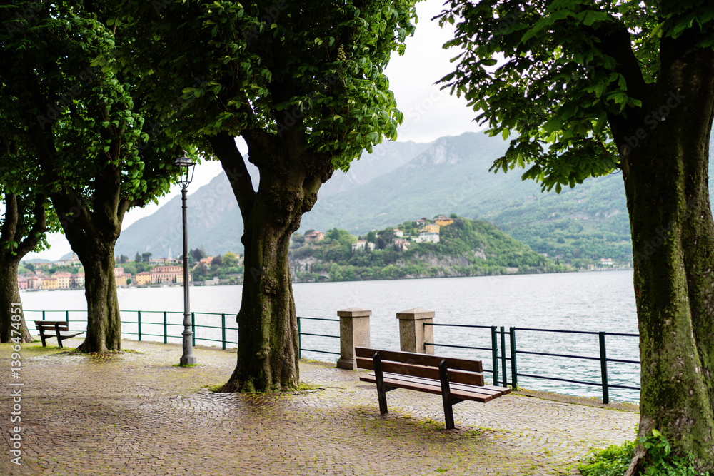 Quiet bench sits under the shade of large trees along lakeside path, offering peaceful view of the distant village and mountains across the water near Lecco at lake Como, Italy Quiet bench sits under the shade of large trees along lakeside path, offering peaceful view of the distant village and mountains across the water near Lecco at lake Como, Italy